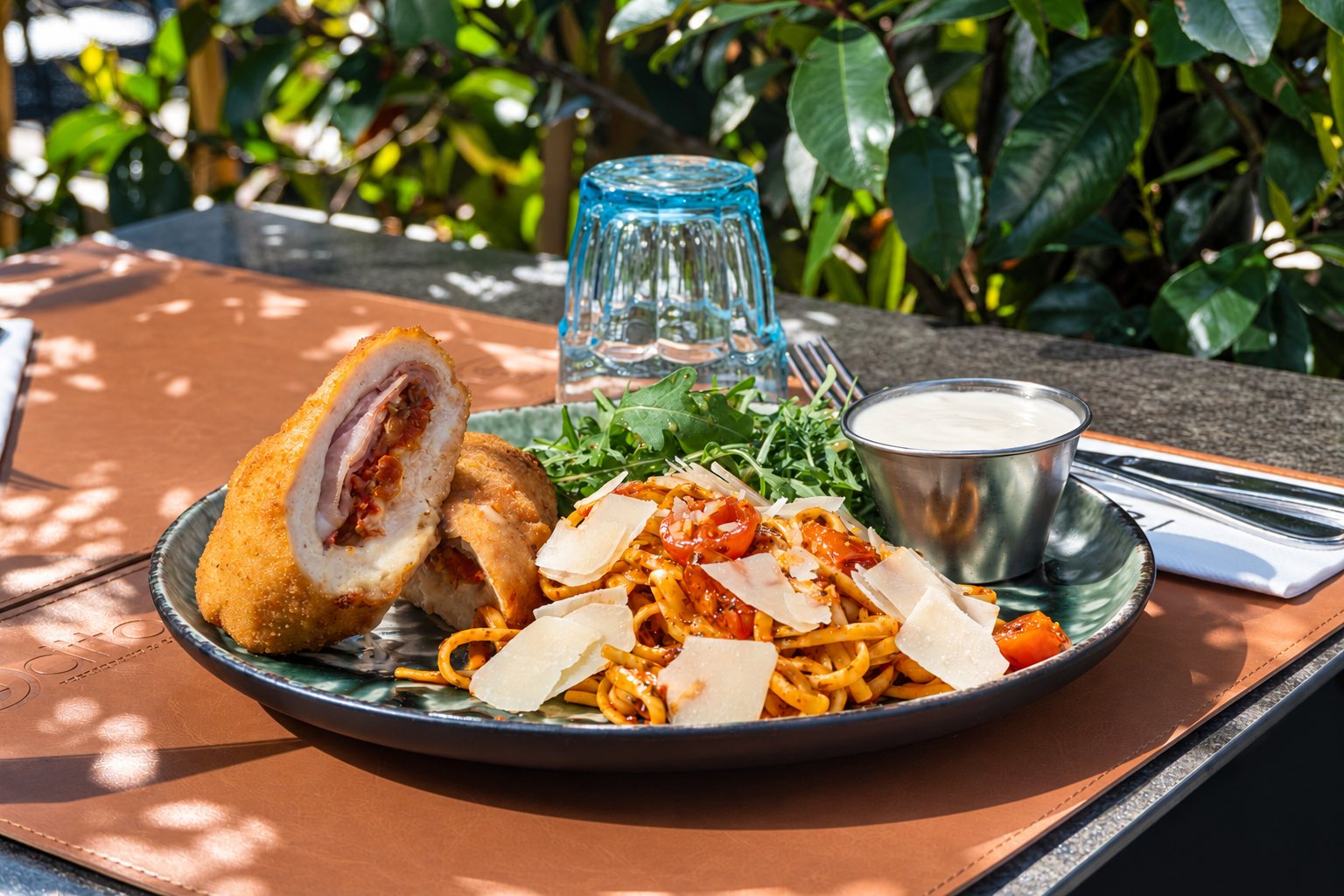 Une assiette de linguine aux tomates cerises et copeaux de parmesan, accompagnée d'une escalope panée farcie, de roquette et d'une sauce blanche, posée sur un set de table en cuir à l'ombre d'un feuillage.