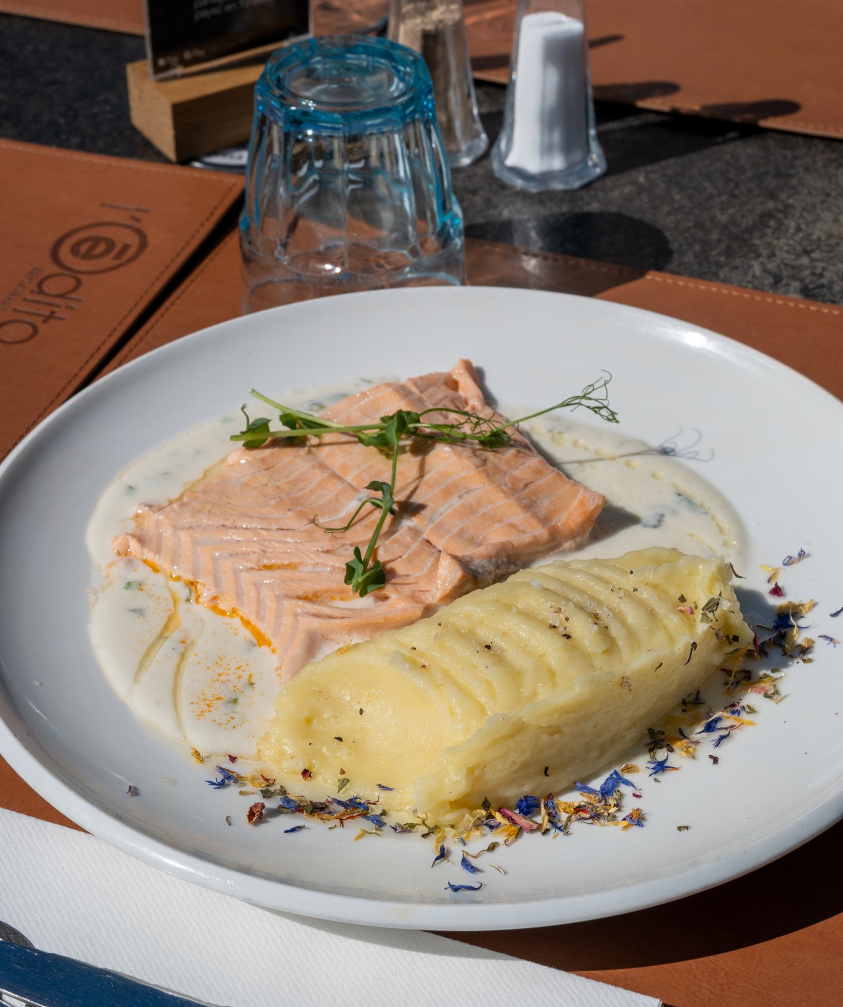 Une assiette blanche garnie d'un filet de saumon nappé de sauce crémeuse, accompagné d'une quenelle de purée et parsemé de pétales de fleurs, posée sur un set de table en cuir sur une terrasse.