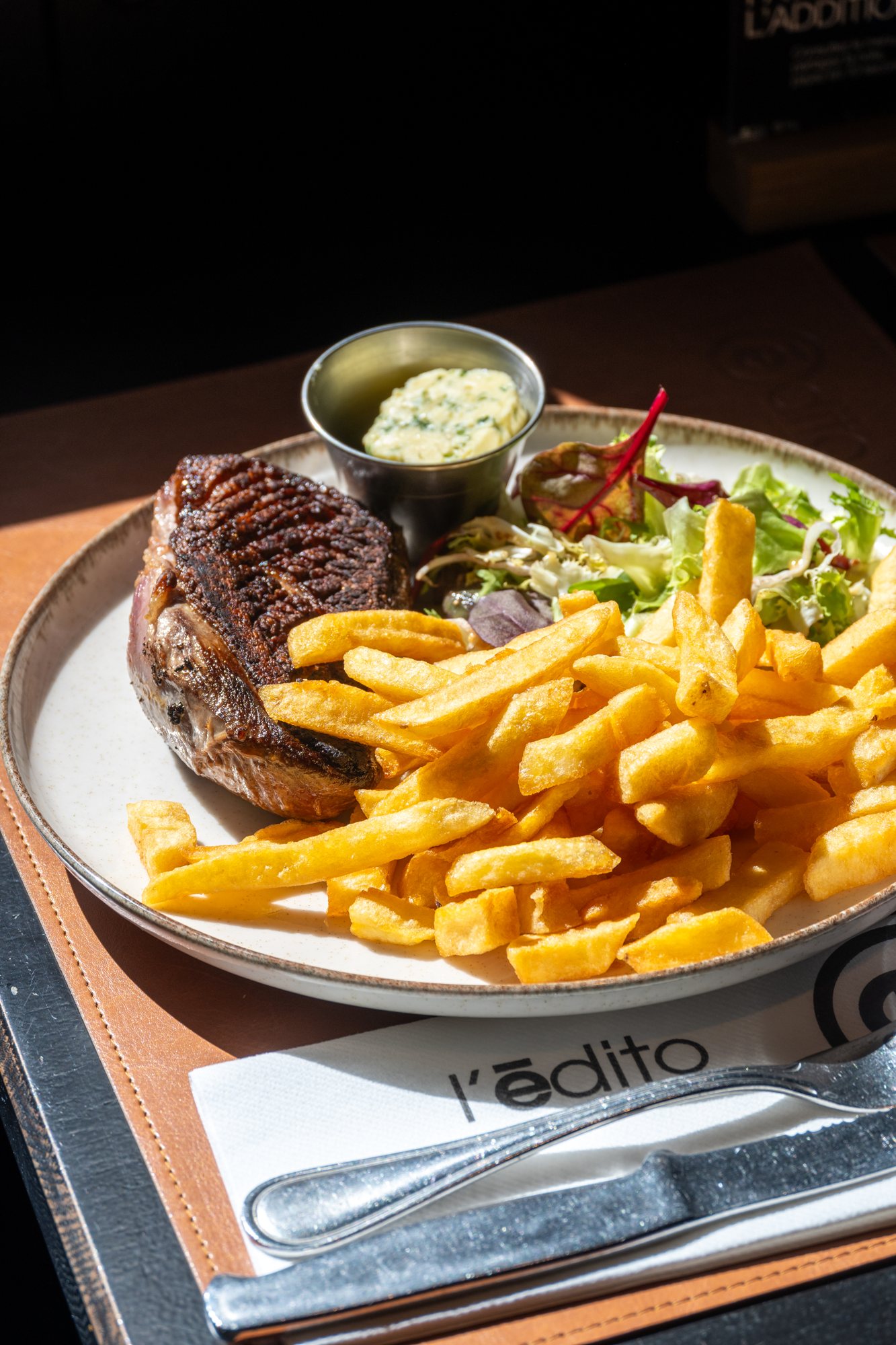 Une assiette de viande grillée accompagnée de frites dorées, de salade verte et d'un petit pot de beurre aux herbes, posée sur un set de table en cuir avec une serviette et des couverts.