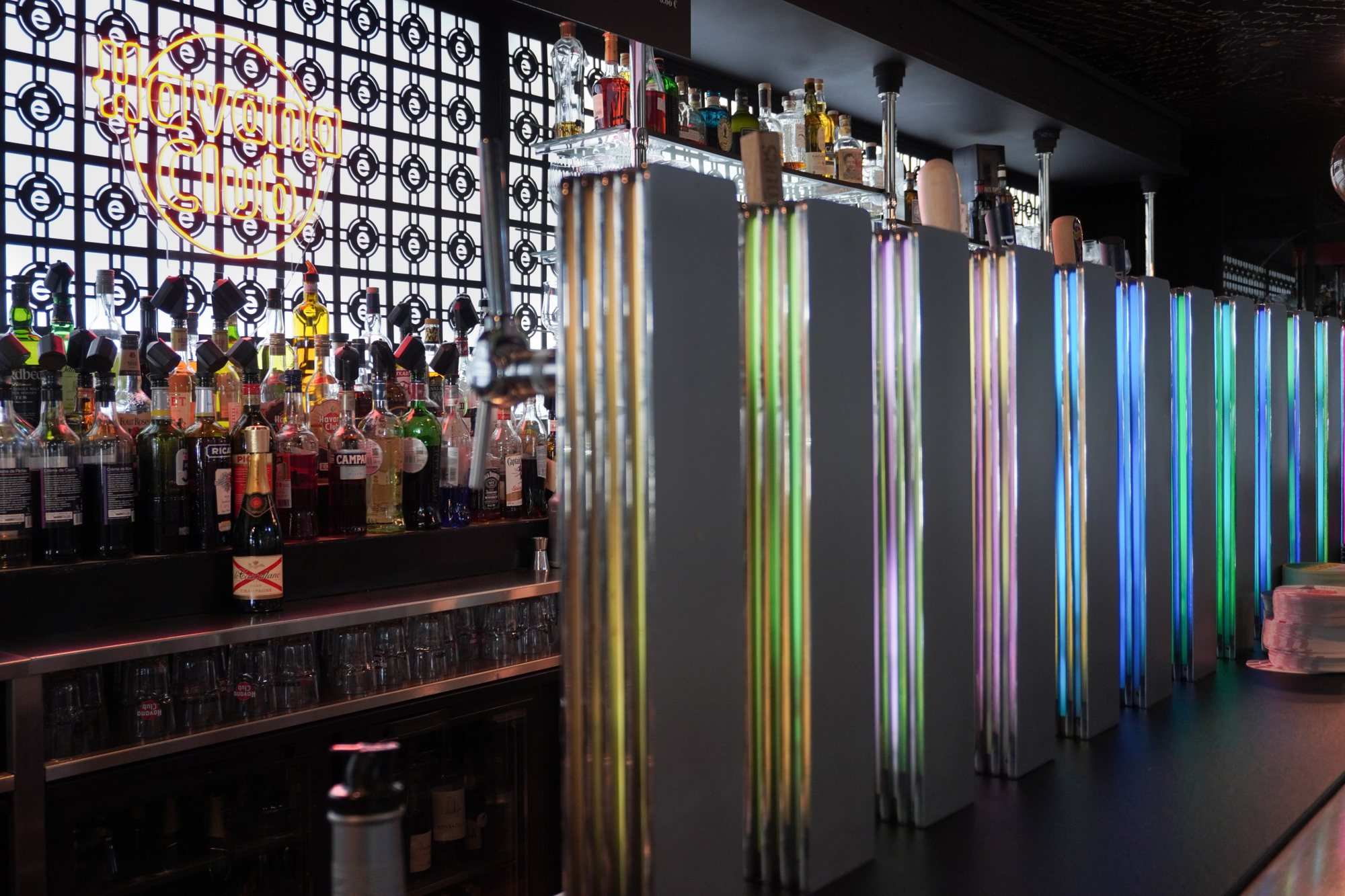 Un comptoir de bar moderne avec une rangée de tireuses à bière éclairées de couleurs vives, des bouteilles d'alcool sur l'étagère et une enseigne néon Havana Club au fond.