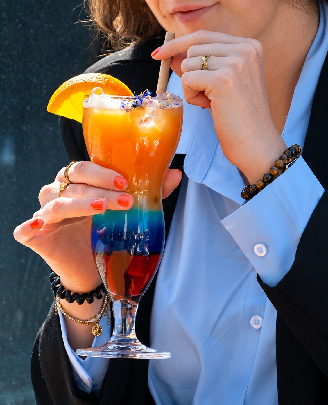 Une femme en chemise bleu clair et veste noire tient un grand verre de cocktail en couches orange, bleu et rouge, décoré d'une tranche d'orange et de fleurs, et boit avec une paille.