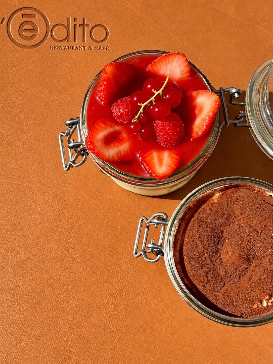 Vue de dessus de deux desserts en bocaux de verre, l'un garni de fraises, framboises et groseilles, l'autre saupoudré de cacao, posés sur un set de table en cuir portant le logo du restaurant.