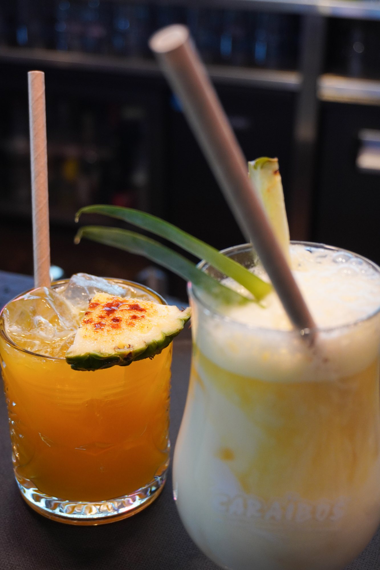 Deux cocktails à l'ananas sur un comptoir de bar : un verre orange avec glaçons et tranche d'ananas caramélisée, et un cocktail crémeux et mousseux avec une paille grise.