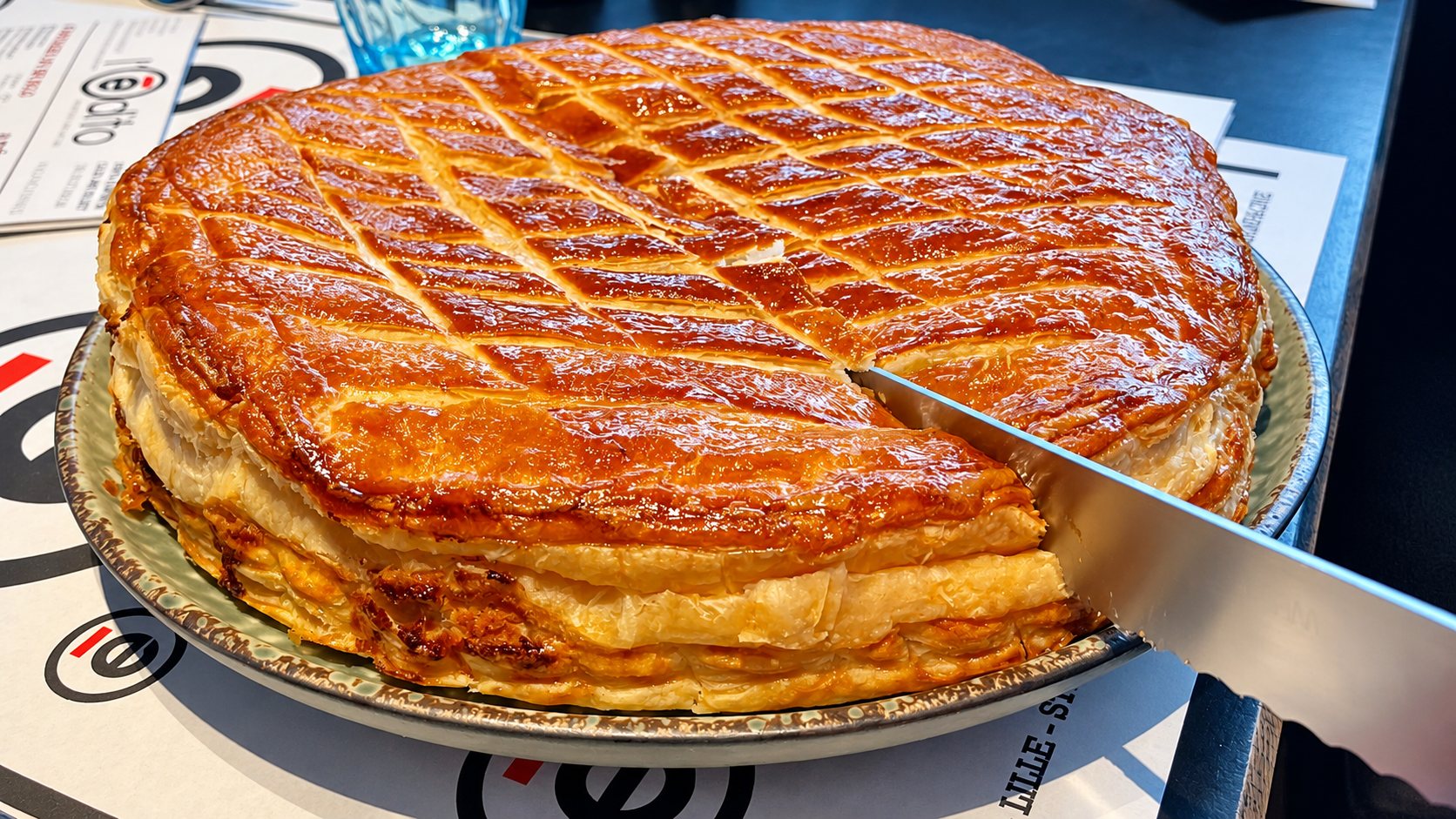 Une galette des rois feuilletée et dorée, posée sur une assiette verte, est entamée par un grand couteau sur une table de restaurant.
