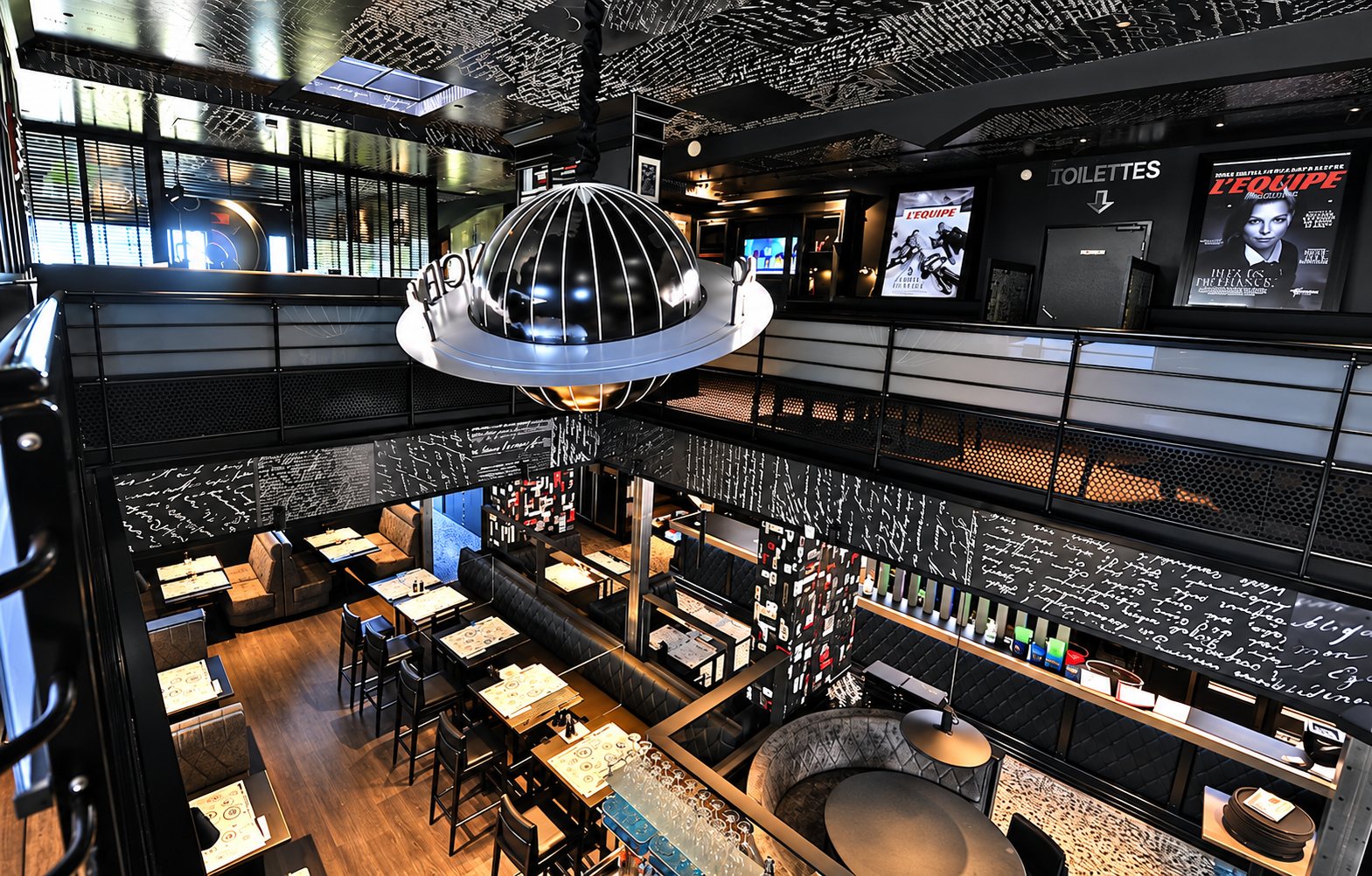 Vue en hauteur d'une salle de restaurant sombre et moderne avec tables, banquettes, mezzanine, grande suspension en forme de planète et affiches de L'Équipe.