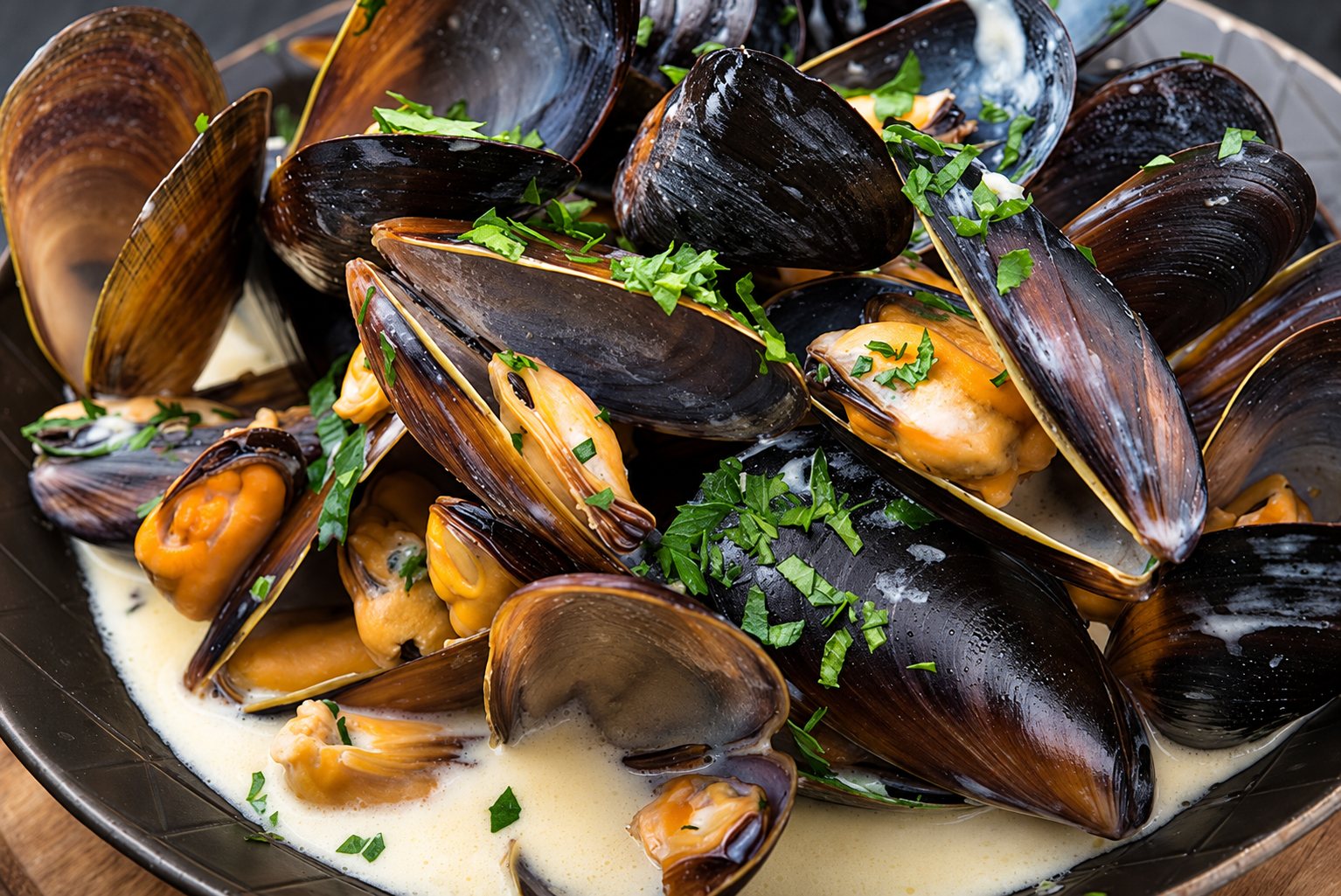 Gros plan sur une assiette creuse de moules ouvertes dans une sauce blanche crémeuse, parsemées de persil haché.