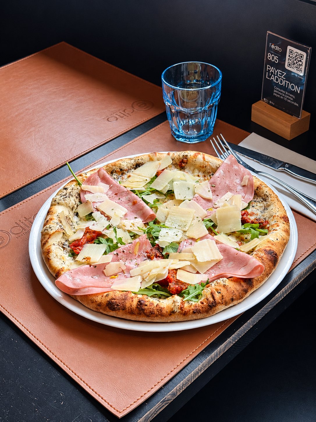 Une pizza garnie de mortadelle, de copeaux de parmesan et de roquette est posée sur un set de table en cuir, à côté d'un verre bleu et de couverts.