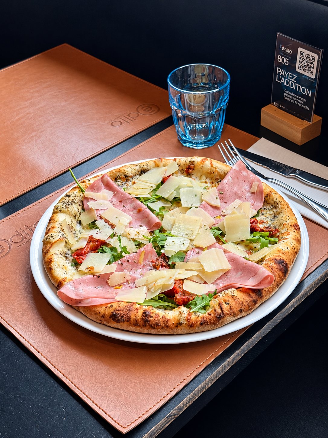 Une pizza garnie de mortadelle, de roquette et de copeaux de parmesan est posée sur un set de table en cuir à côté d'un verre bleu et de couverts.
