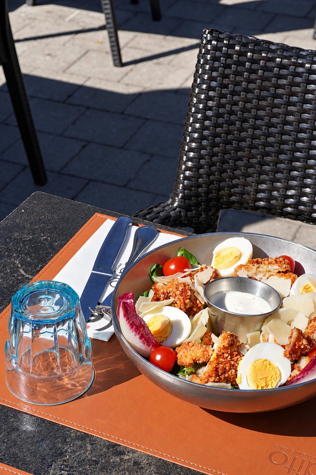 Un grand bol métallique de salade César avec poulet pané, œufs durs, tomates cerises et copeaux de parmesan, posé sur un set de table en cuir orange à côté d'un verre bleu retourné, sur une table de terrasse.