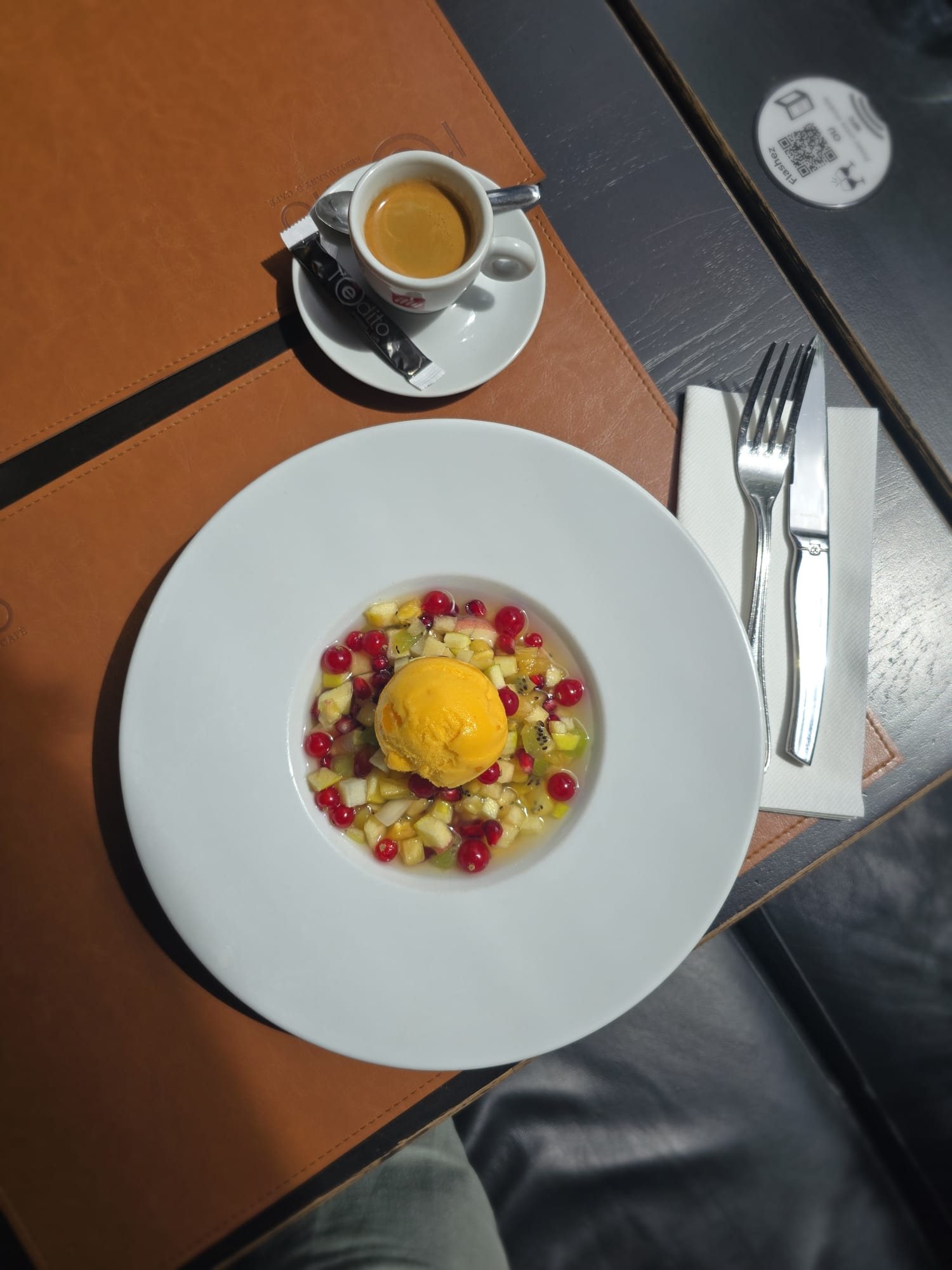 Vue de dessus d'une assiette creuse blanche contenant une salade de fruits frais et une boule de sorbet orange, posée sur un set de table en cuir à côté d'un expresso.