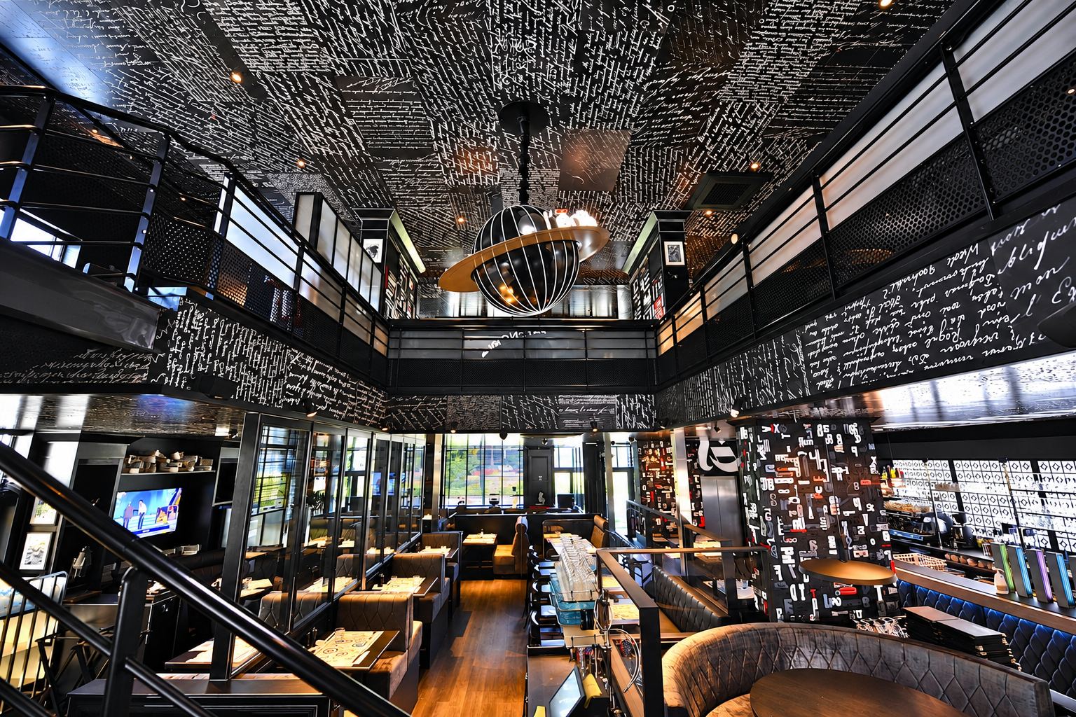Vaste salle de restaurant sur deux niveaux, aux murs et au plafond noirs couverts d'écriture manuscrite blanche, avec banquettes capitonnées et suspension en forme de globe.