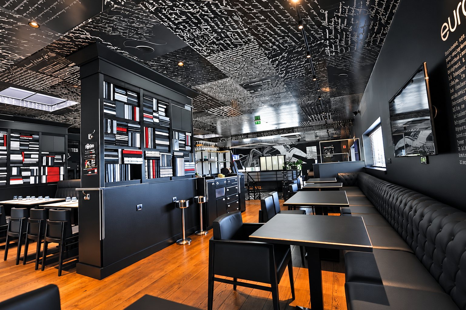 Intérieur d'un restaurant aux murs et au plafond noirs couverts d'écritures blanches, avec banquette capitonnée, tables sombres et bibliothèque centrale.