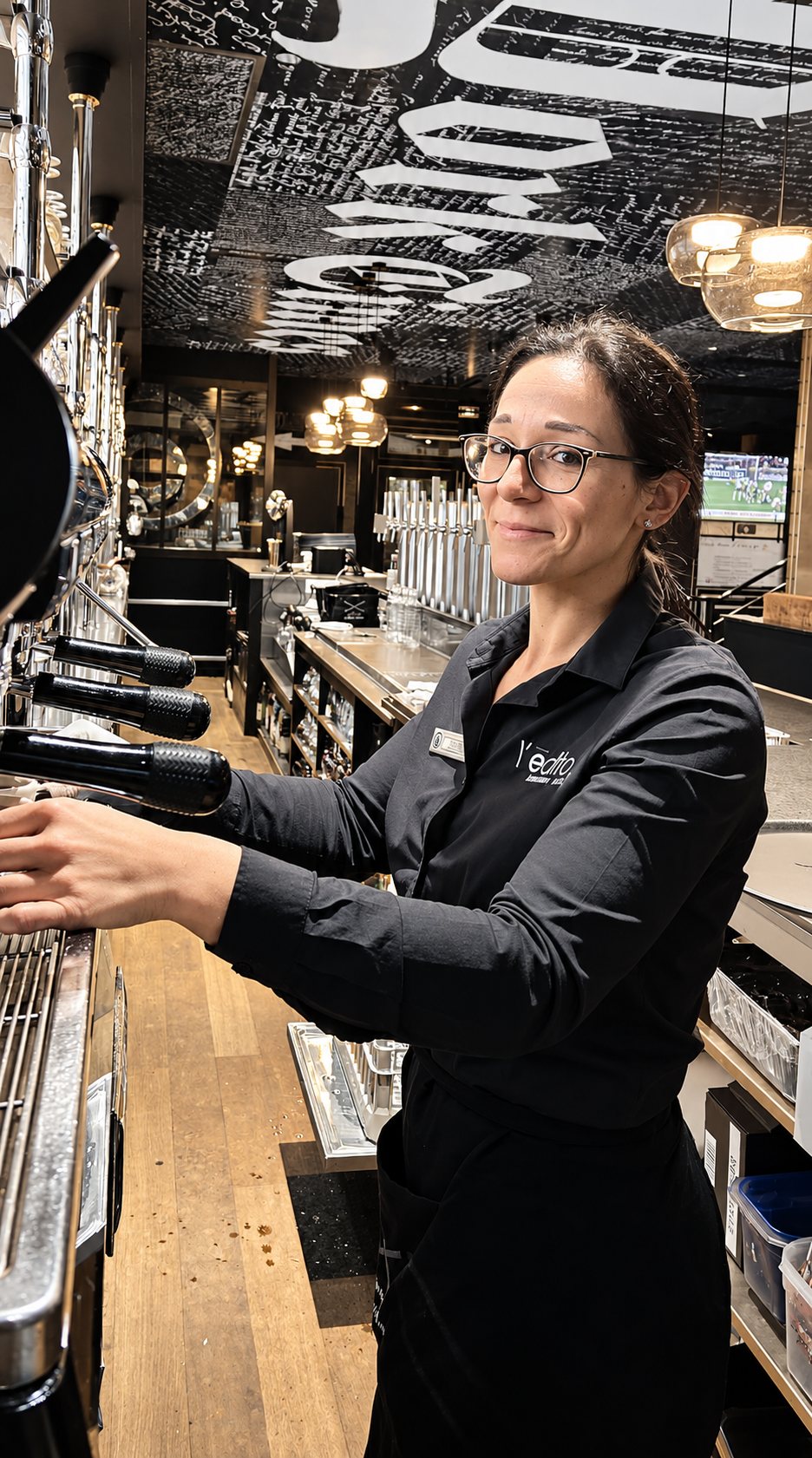 Une serveuse à lunettes sourit derrière le comptoir d'un bar, les mains posées sur une machine à café, en noir et blanc.