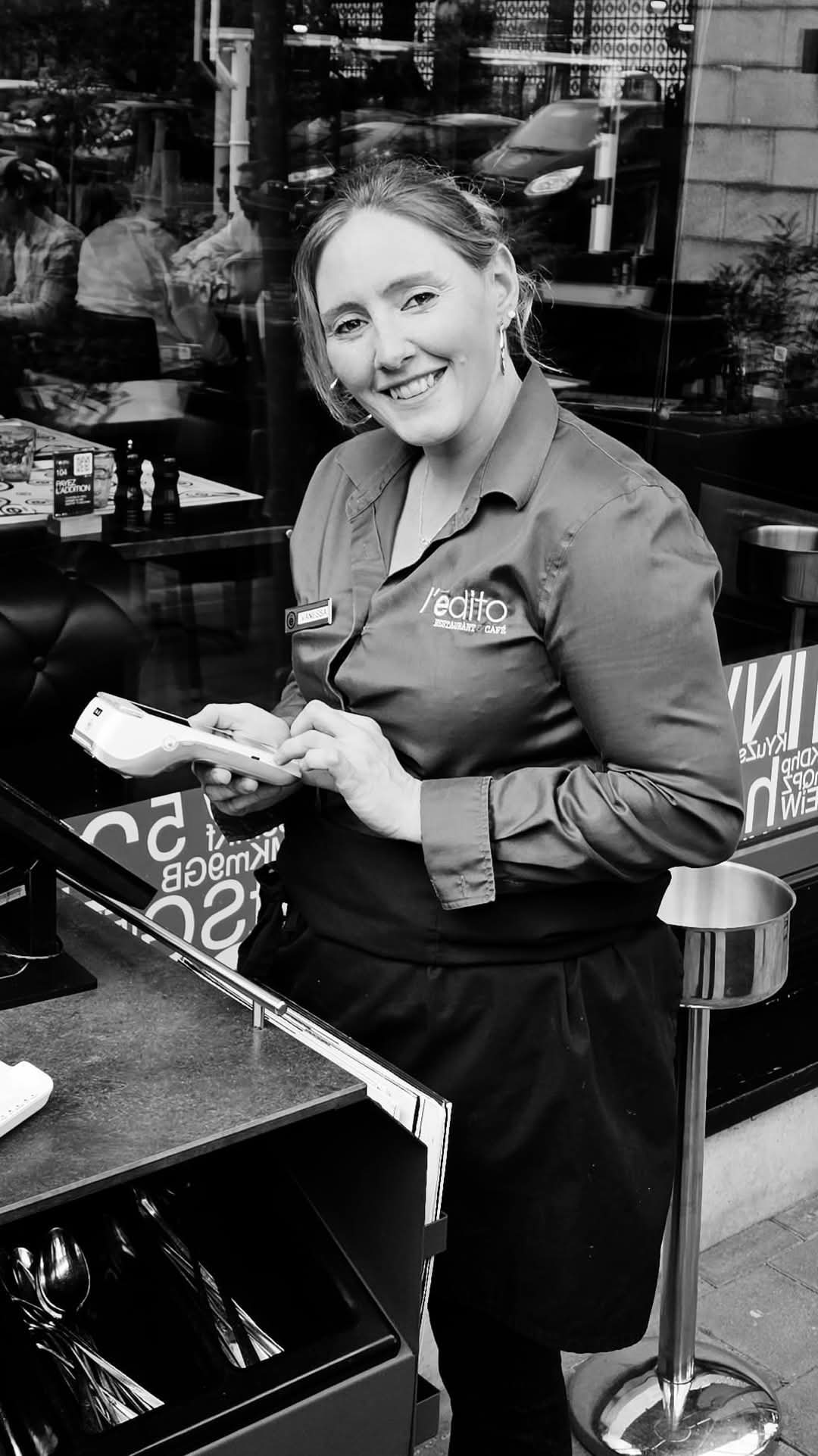 Une serveuse en uniforme sourit en tenant un terminal de paiement à côté d'un meuble à couverts sur la terrasse d'un restaurant, en noir et blanc.
