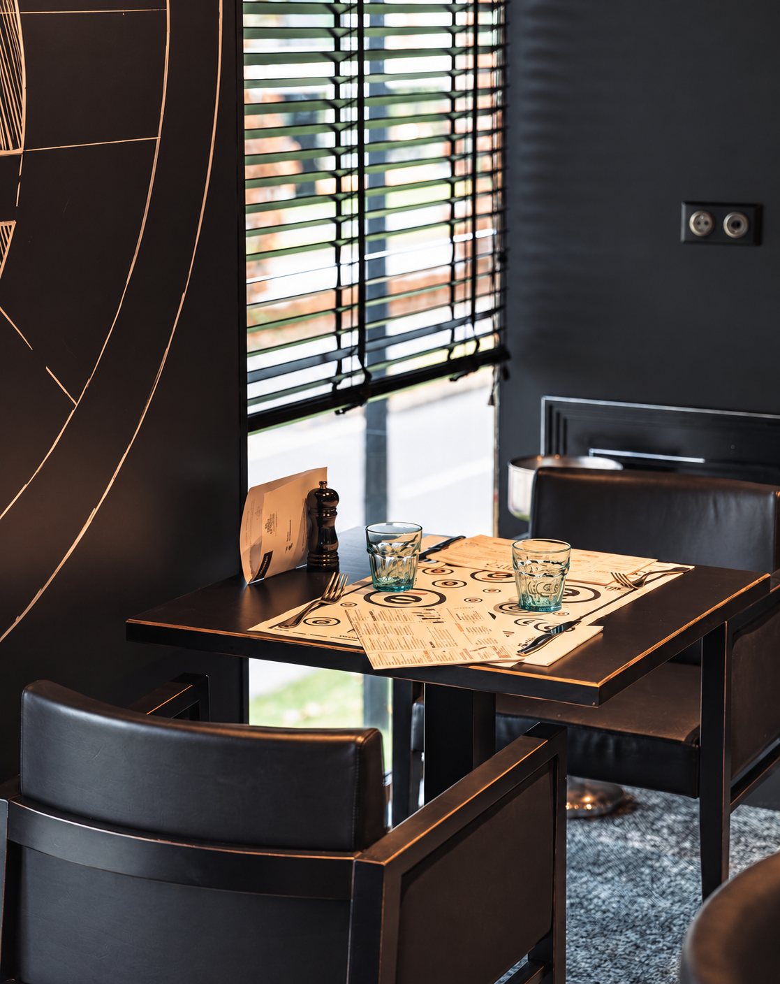 Une petite table carrée avec set de table imprimé, verres bleutés et menus, entourée de fauteuils en cuir noir près d'un store vénitien.