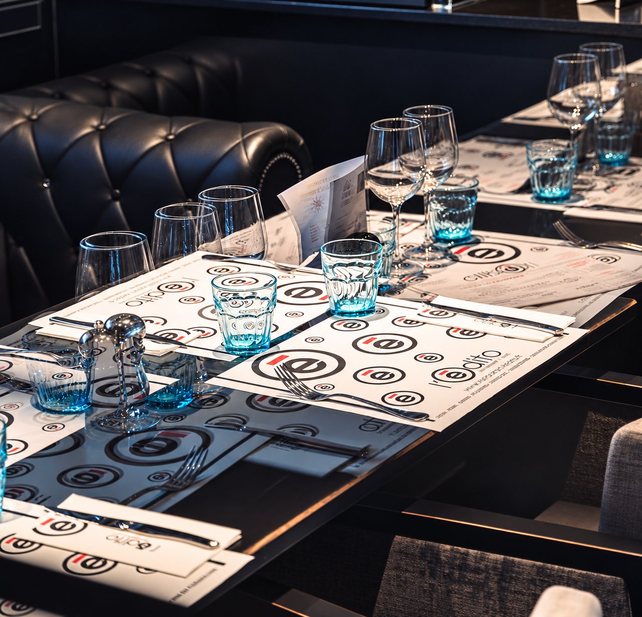 Une table de restaurant dressée avec des sets de table imprimés au logo, des verres bleutés et des couverts, devant une banquette en cuir sombre.