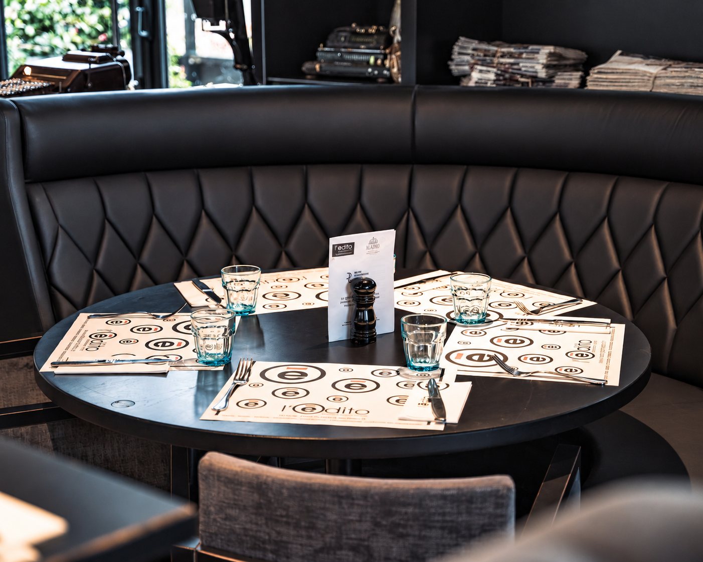 Une table ronde noire dressée avec quatre sets de table imprimés, des verres et des couverts, devant une banquette en cuir capitonné sombre.