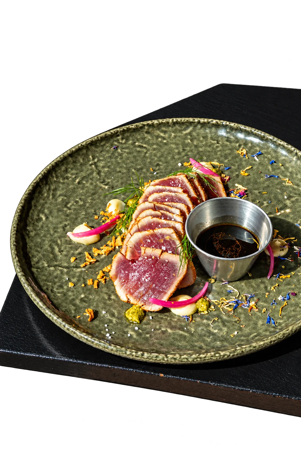 Tranches de thon mi-cuit disposées sur une assiette en céramique verte, avec oignon rouge mariné, aneth, fleurs séchées et un petit pot de sauce soja.