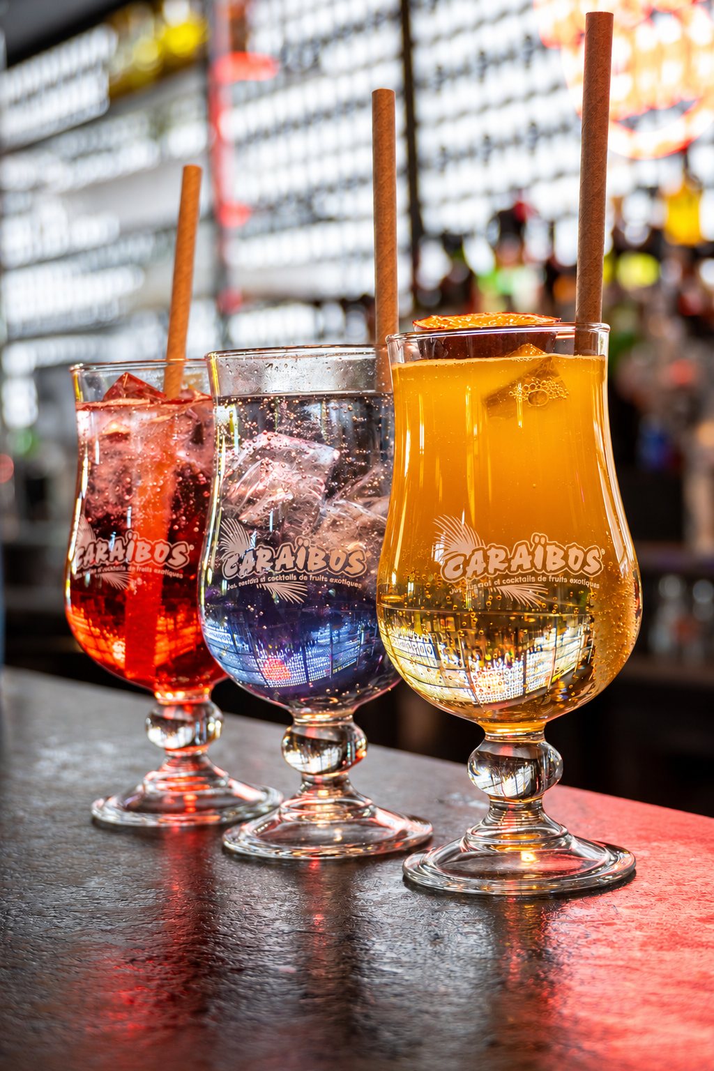 Trois verres à pied ornés du logo Caraibos, remplis de cocktails rouge, bleu et orange avec des pailles, posés sur un comptoir de bar.